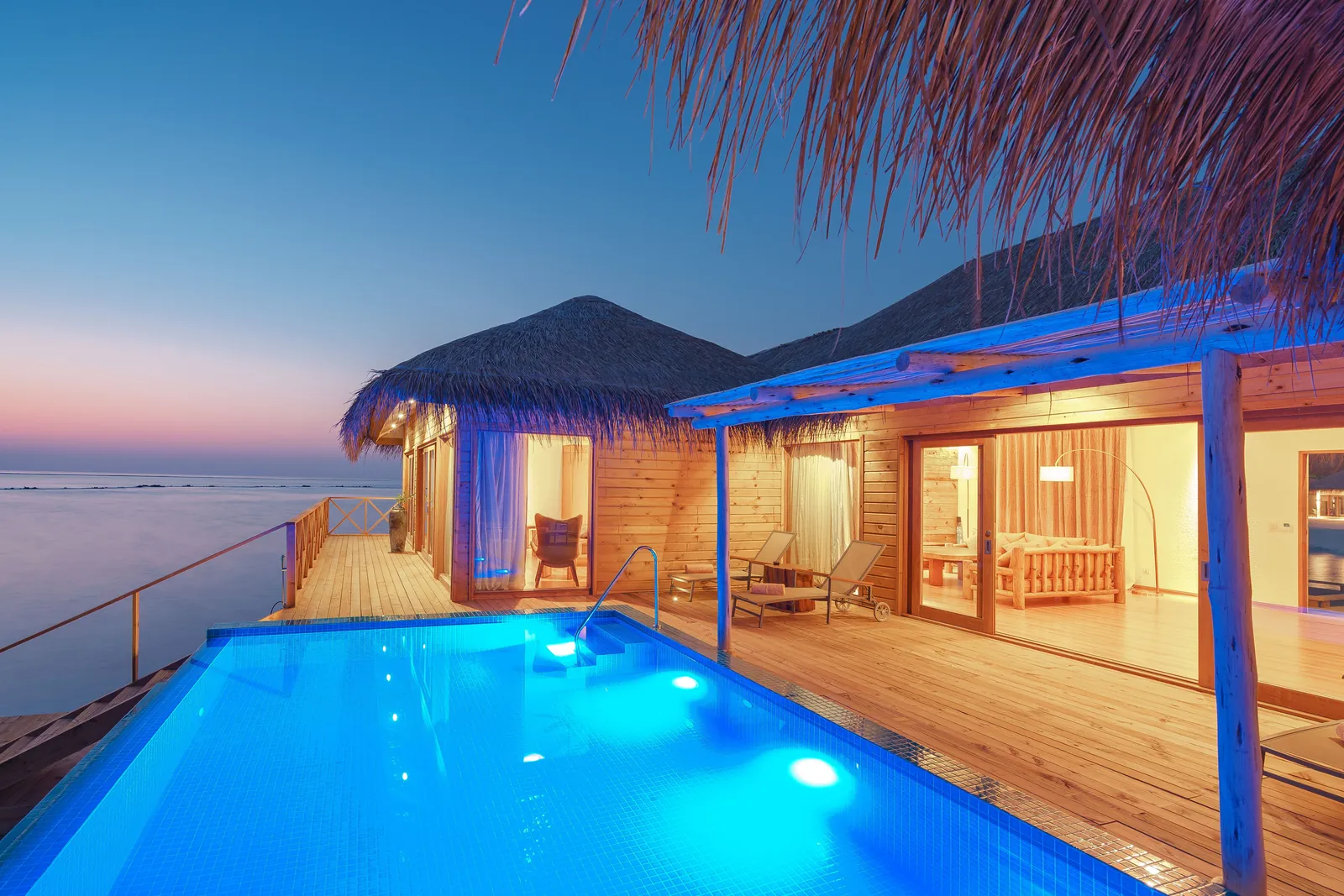 Private overwater villa with a glowing infinity-edge plunge pool at dusk, thatched roofs and warm lantern light against a fading tropical sky.